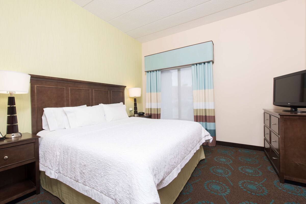 A hotel room with a large bed, two bedside tables with lamps, a television on a dresser, and a window with curtains.
