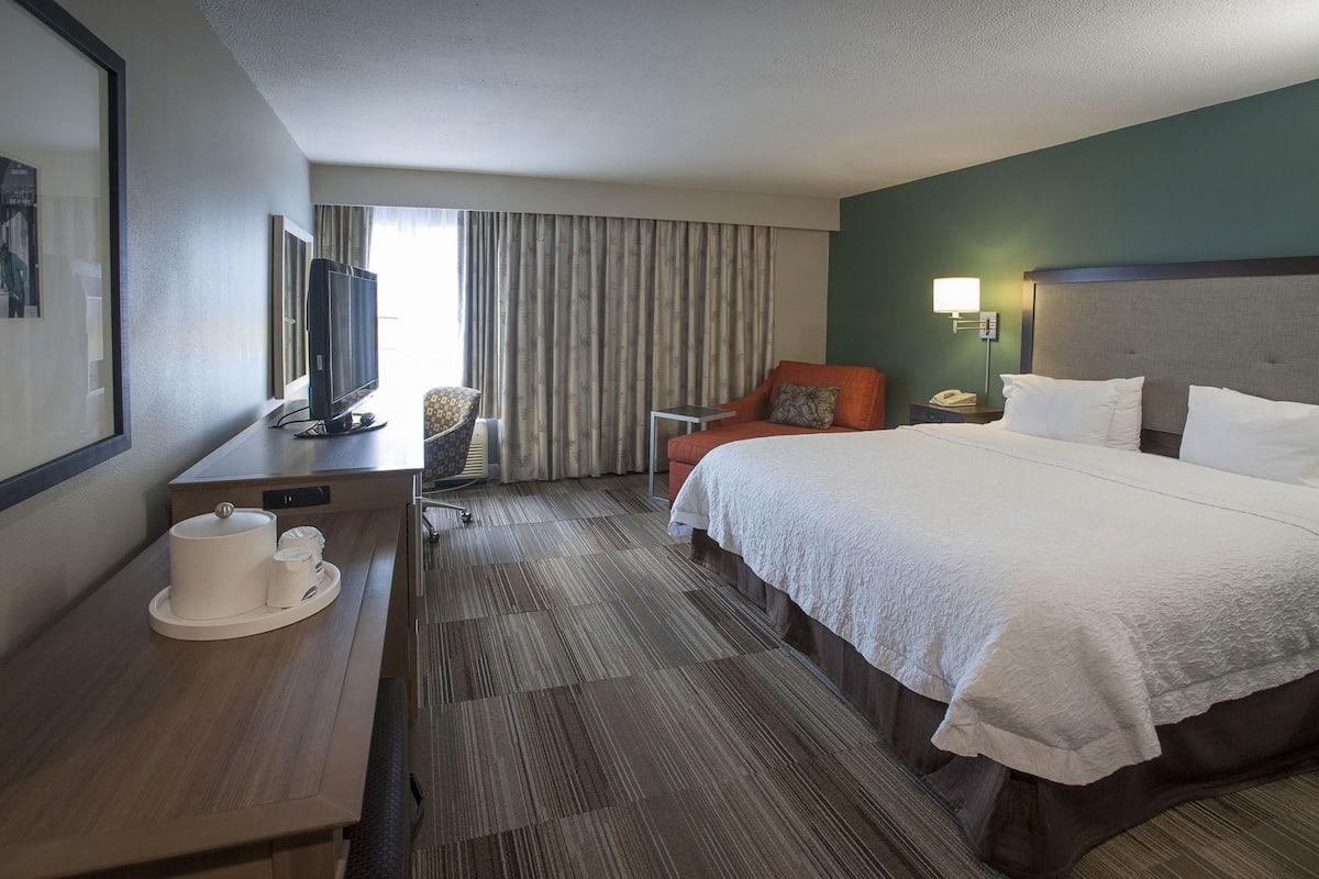 A hotel room with a bed, a desk with a television, a chair, a lamp, and a window with curtains.