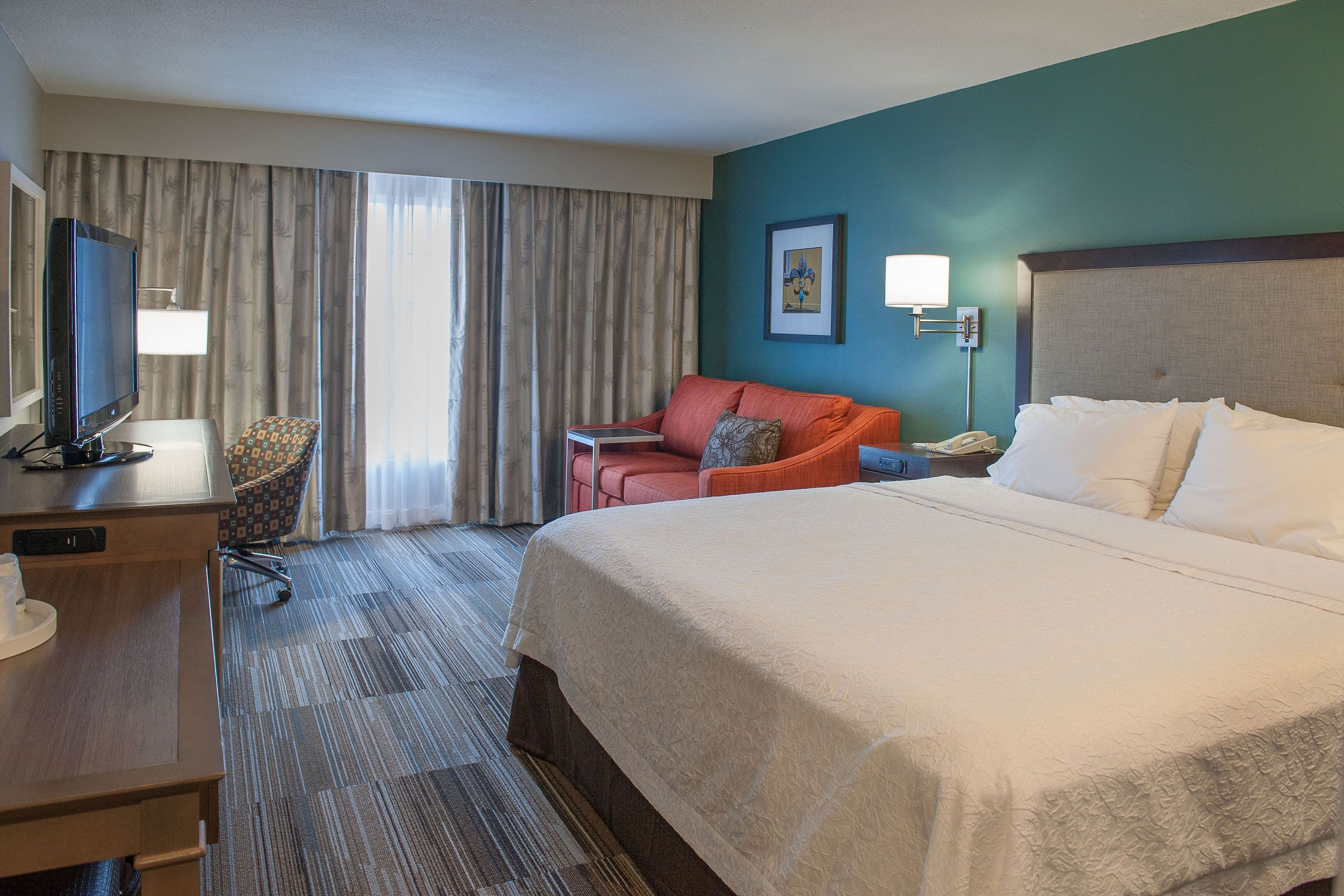 A hotel room with a large bed, a desk with a television, a chair, and a desk lamp.