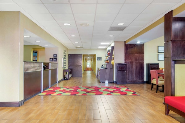 A hotel lobby with a wooden floor, a reception desk, a waiting area with chairs, and a hallway leading to another section.