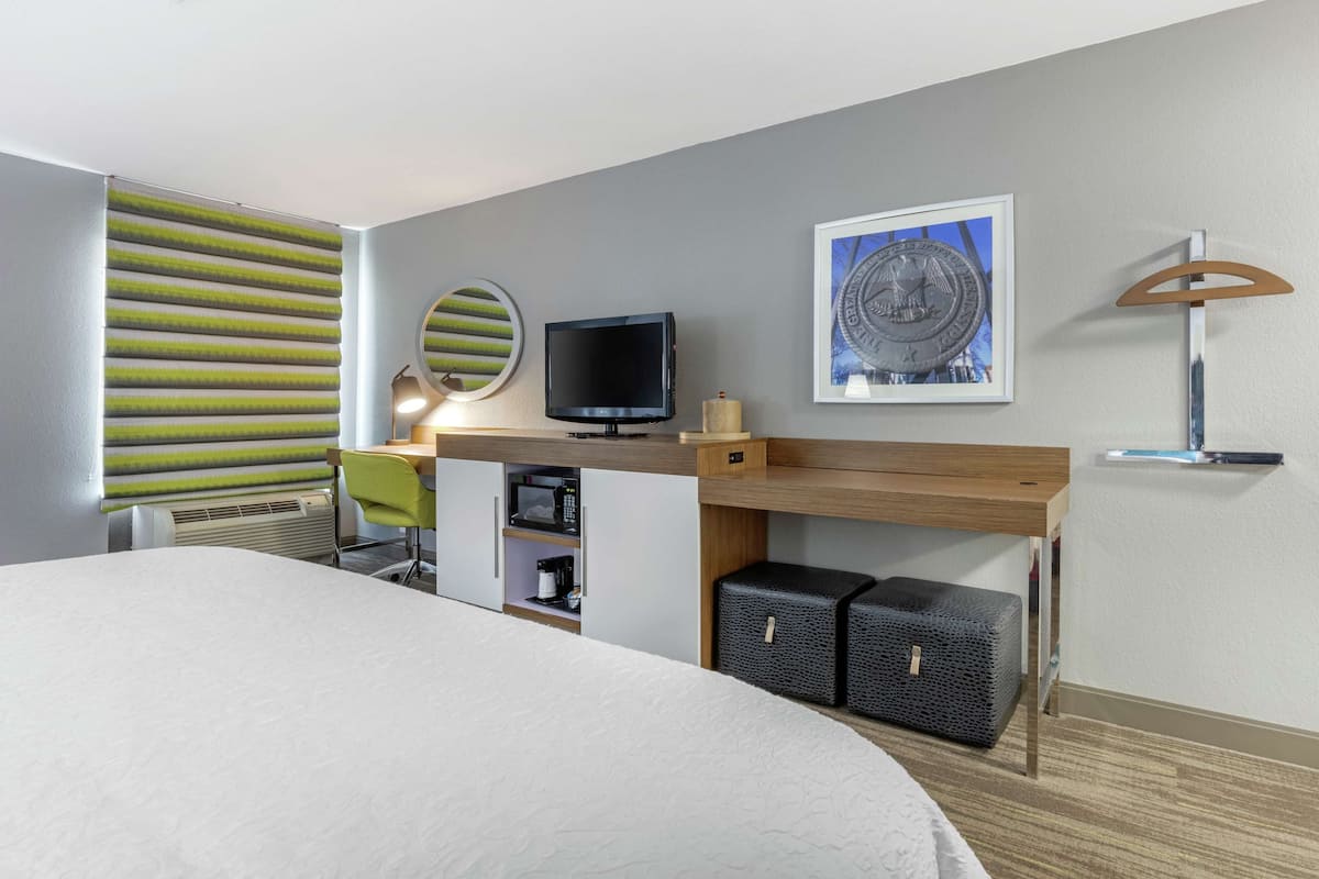 A modern hotel room with a large bed, a desk with a flat-screen TV, a microwave, and storage bins.