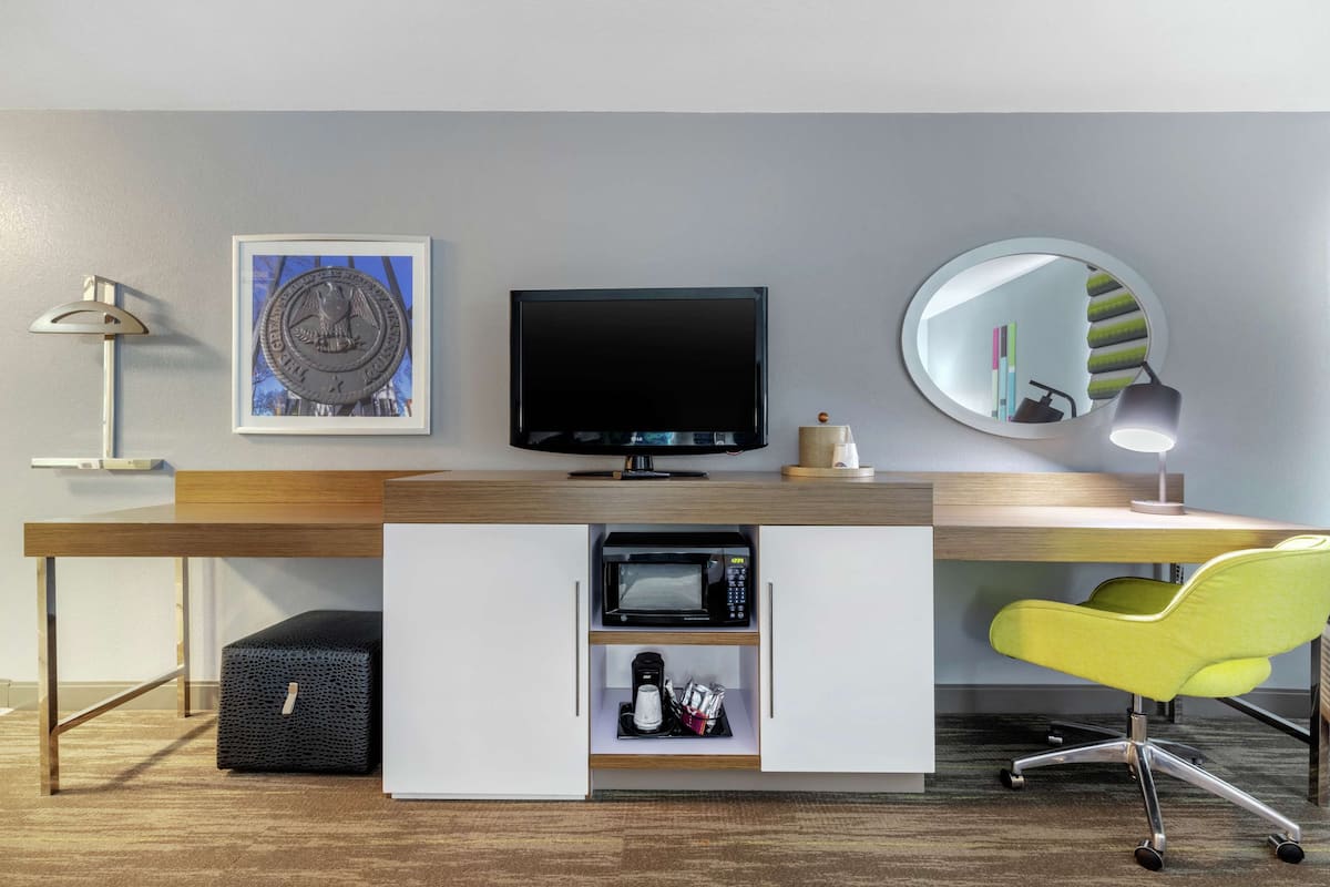 A modern office space with a desk, a television, a microwave, a yellow chair, a lamp, and a framed picture on the wall.