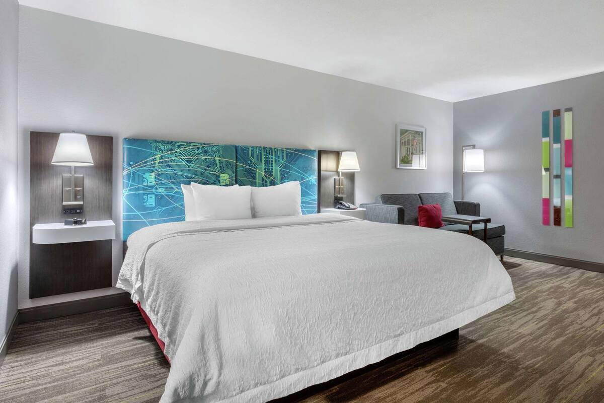 A hotel room with a large bed, bedside lamps, a desk with a chair, a small table, and a wall with a colorful abstract design.