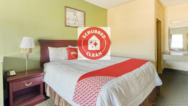 A hotel room with a bed, a nightstand, a lamp, a painting on the wall, and a bathroom visible through an open door.