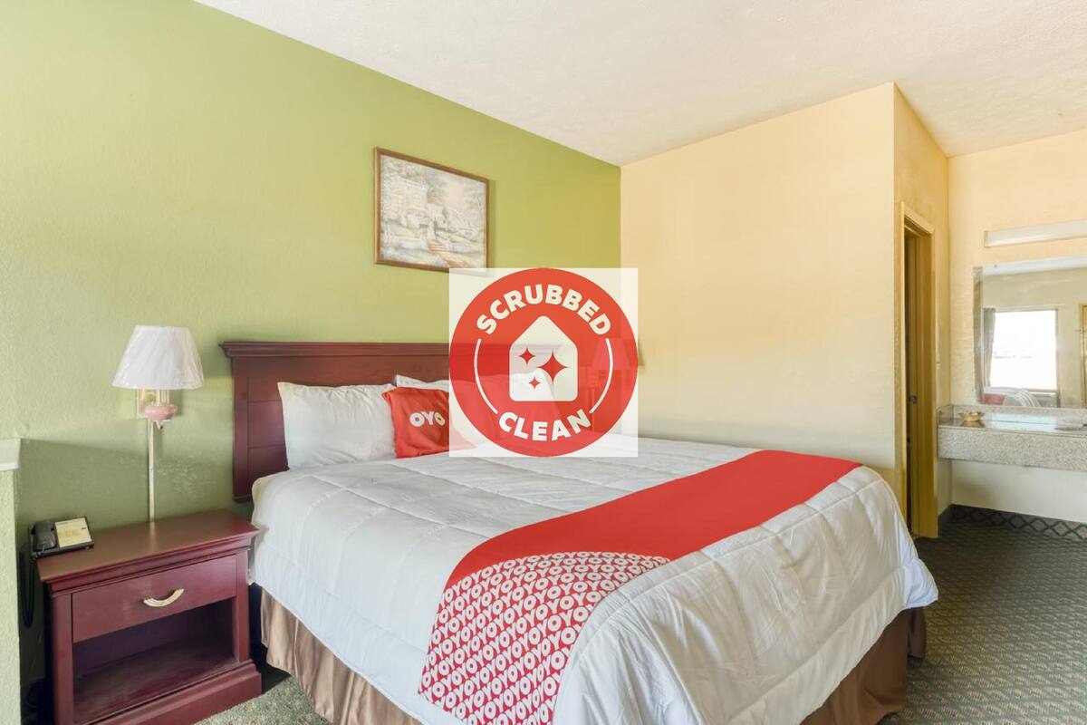A hotel room with a bed, a nightstand, a lamp, a painting on the wall, and a bathroom visible through an open door.
