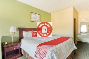 Una habitación de hotel con una cama, una mesita de noche, una lámpara, un cuadro en la pared y un baño visible a través de una puerta abierta.