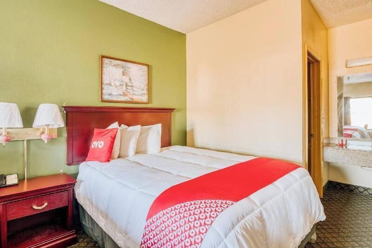 A hotel room with a bed, a nightstand, a lamp, a painting on the wall, and a bathroom visible through an open door.