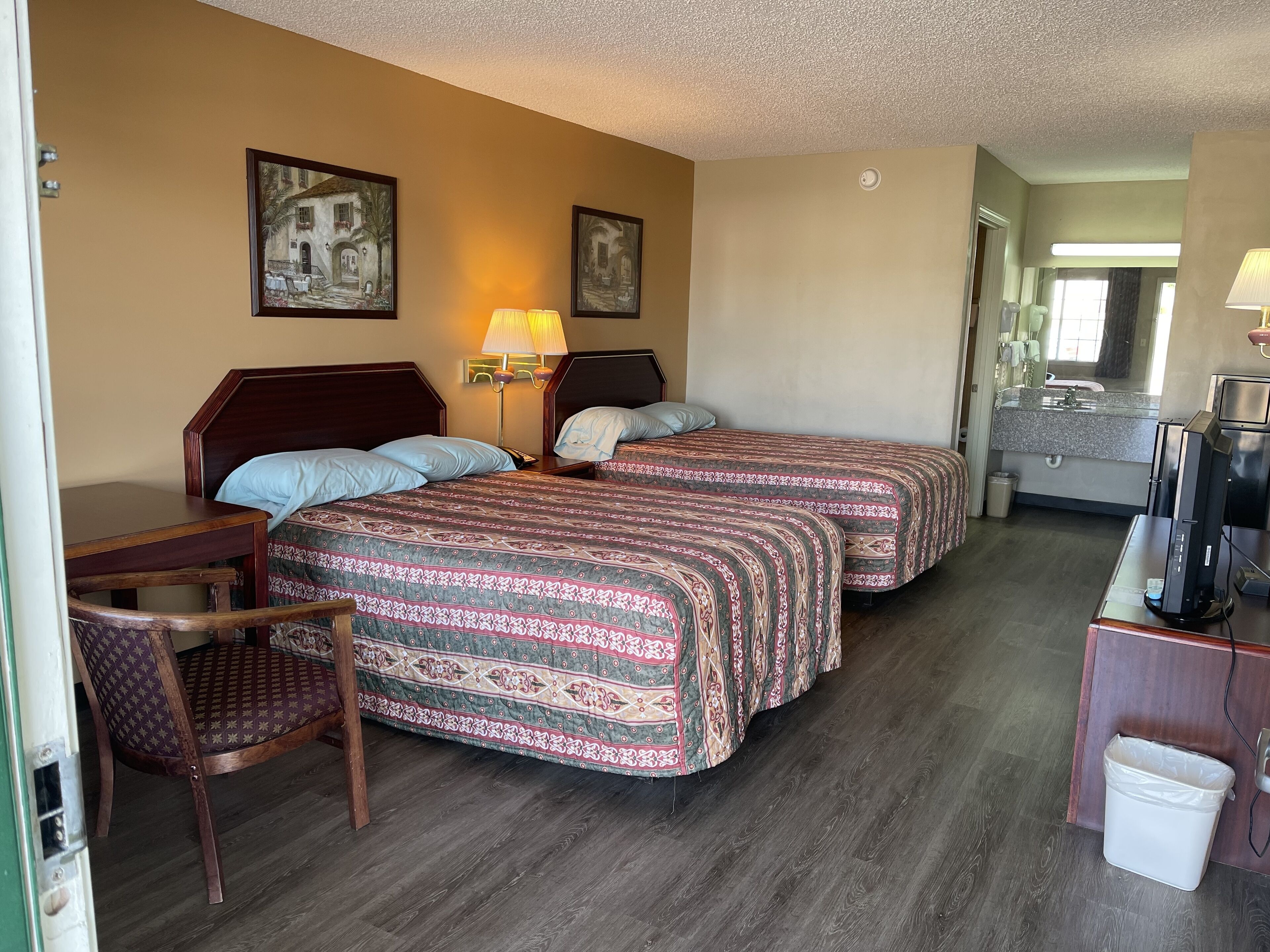 A hotel room with two beds, a desk, a chair, a TV, and a bathroom visible through an open door.