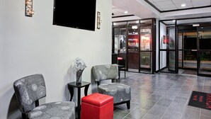 A lobby with two grey upholstered chairs, a red ottoman, a flat-screen TV, and a glass door entrance. - Ramada by Wyndham Pearl/Jackson Airport (Pearl)