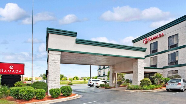 A Ramada hotel with a covered parking area, a flagpole with the American flag, and a signboard. - Ramada by Wyndham Pearl/Jackson Airport (Pearl)