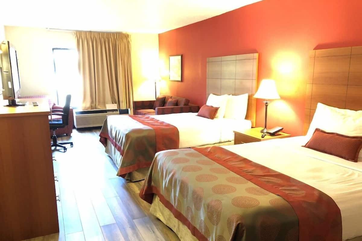 Habitación de hotel con dos camas, un escritorio, una silla, una lámpara y un ventanal con cortinas.