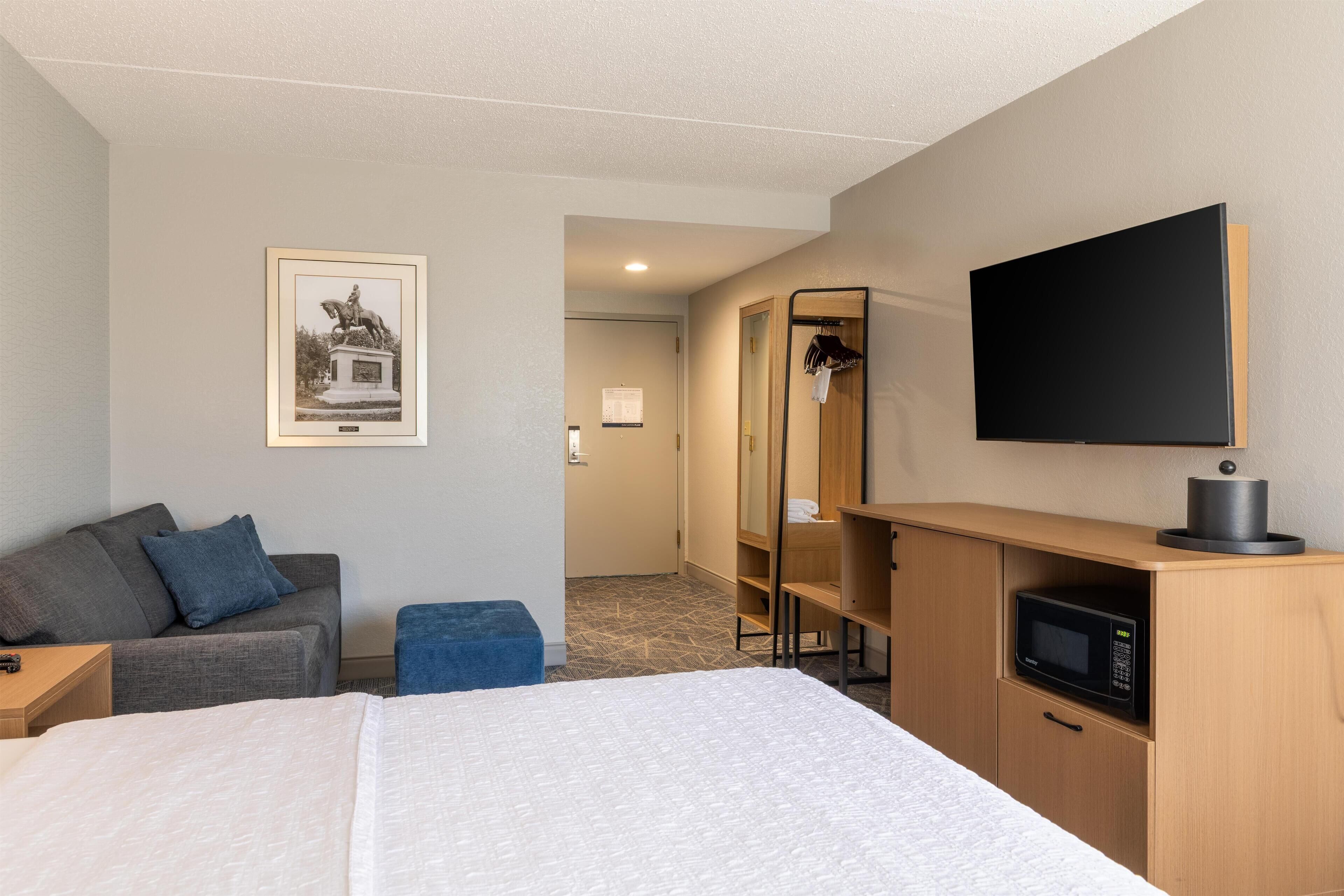 A hotel room with a bed, a gray sofa, a small table, a TV, a microwave, a wooden cabinet, and a framed picture on the wall.