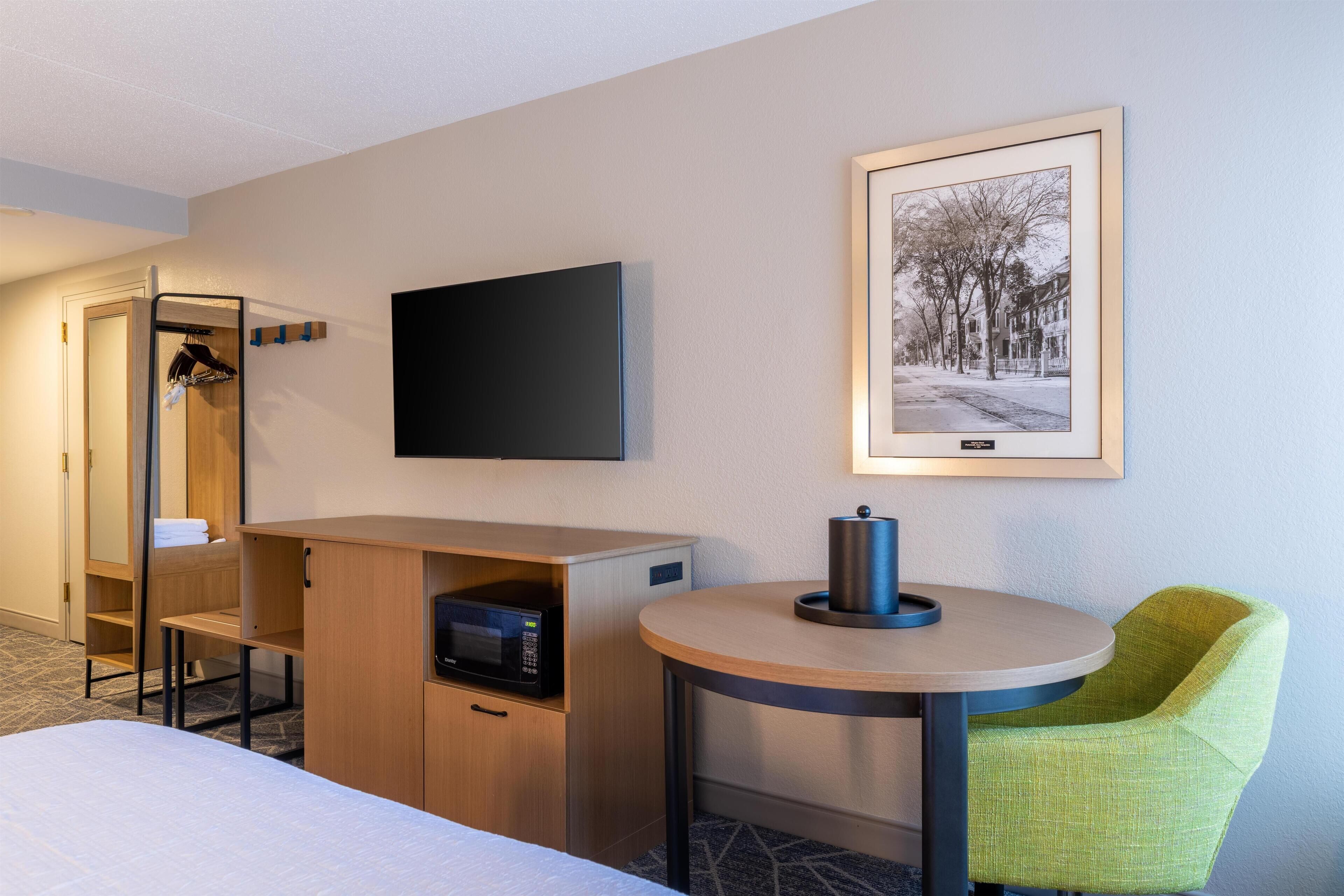A hotel room with a bed, a desk with a microwave, a chair, a TV, and a framed picture on the wall.