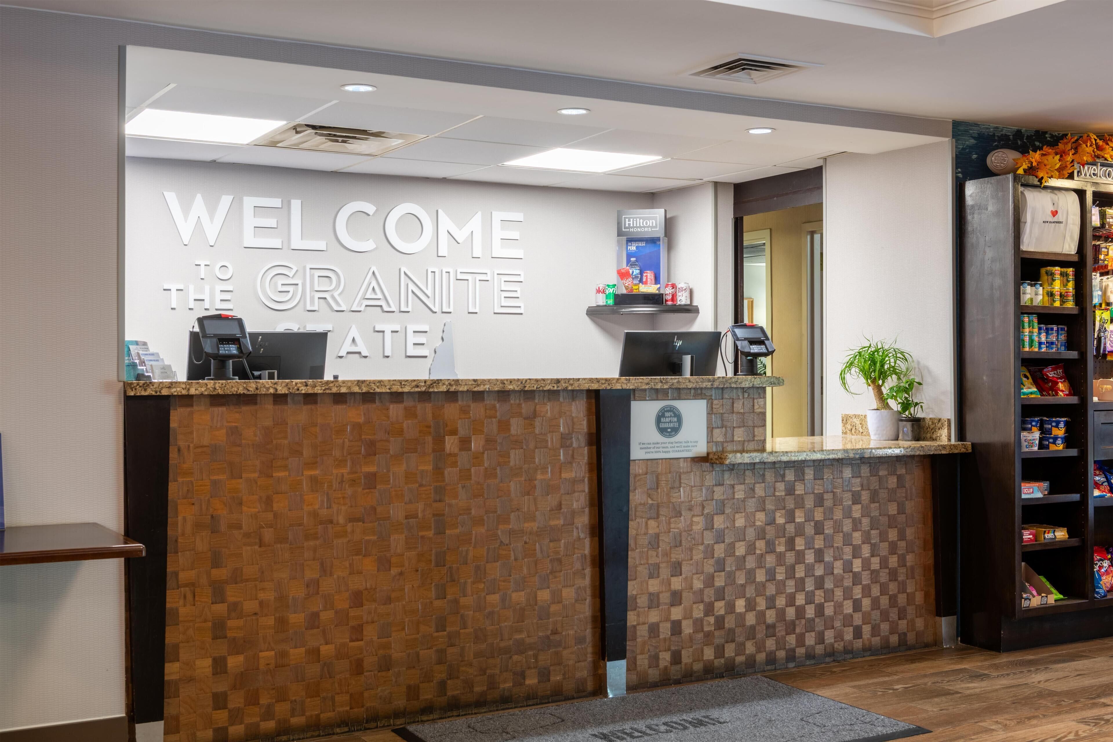 A hotel reception desk with a welcome sign, a coffee machine, a plant, and a vending machine.
