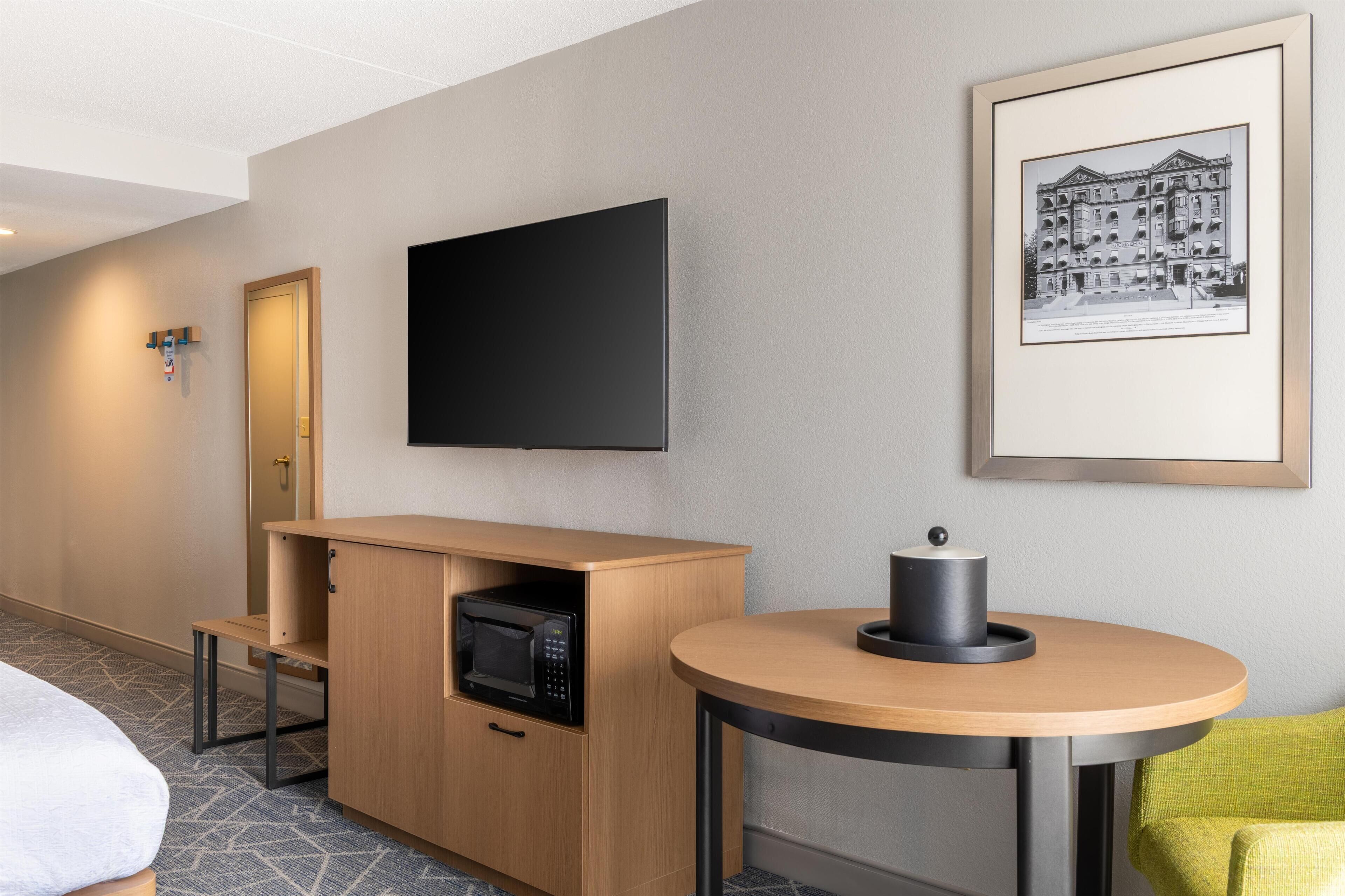 Una habitación de hotel con una cama, un armario de madera con un microondas, una mesa redonda con un objeto negro, un cuadro en la pared y una puerta con manija.