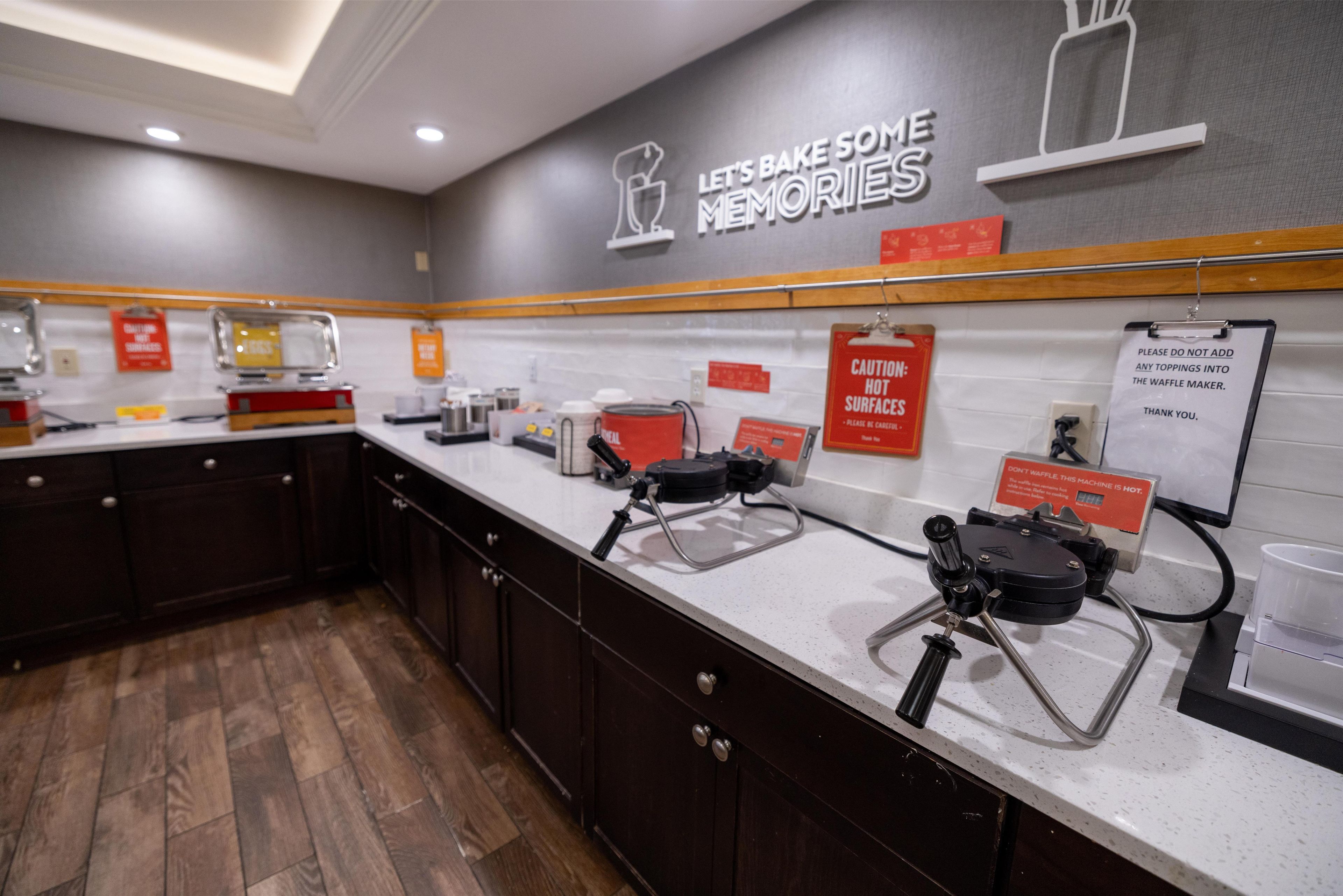 A kitchen area with a sign that reads "Let's Bake Some Memories." There are waffle makers on the counter, and caution signs about hot surfaces and not adding toppings to the waffle maker.