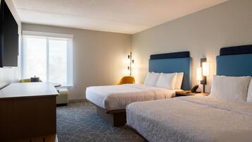 A hotel room with two beds, a desk, a chair, a lamp, and a window.