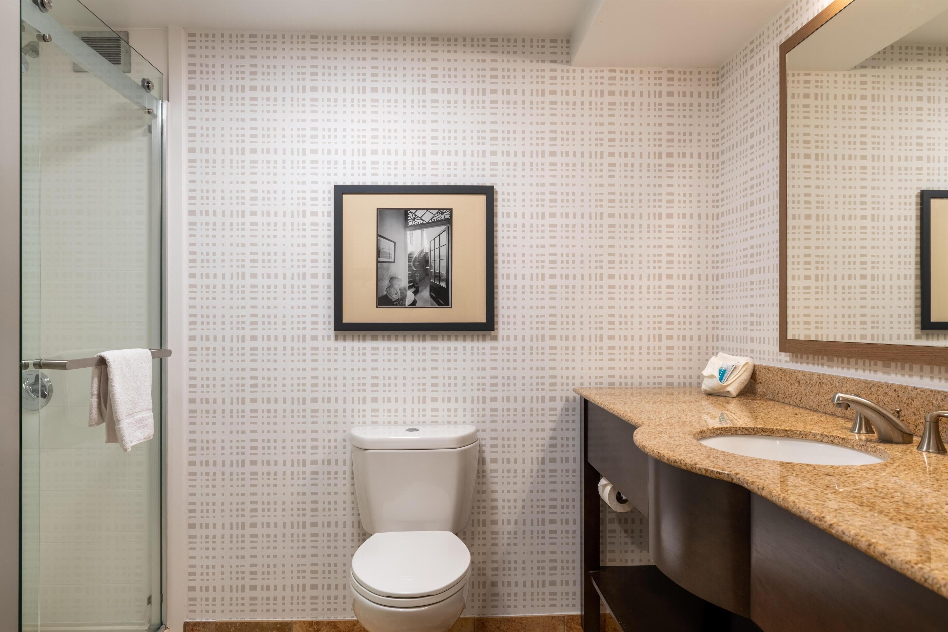 A bathroom with a toilet, a sink, and a shower. There is a framed picture on the wall.
