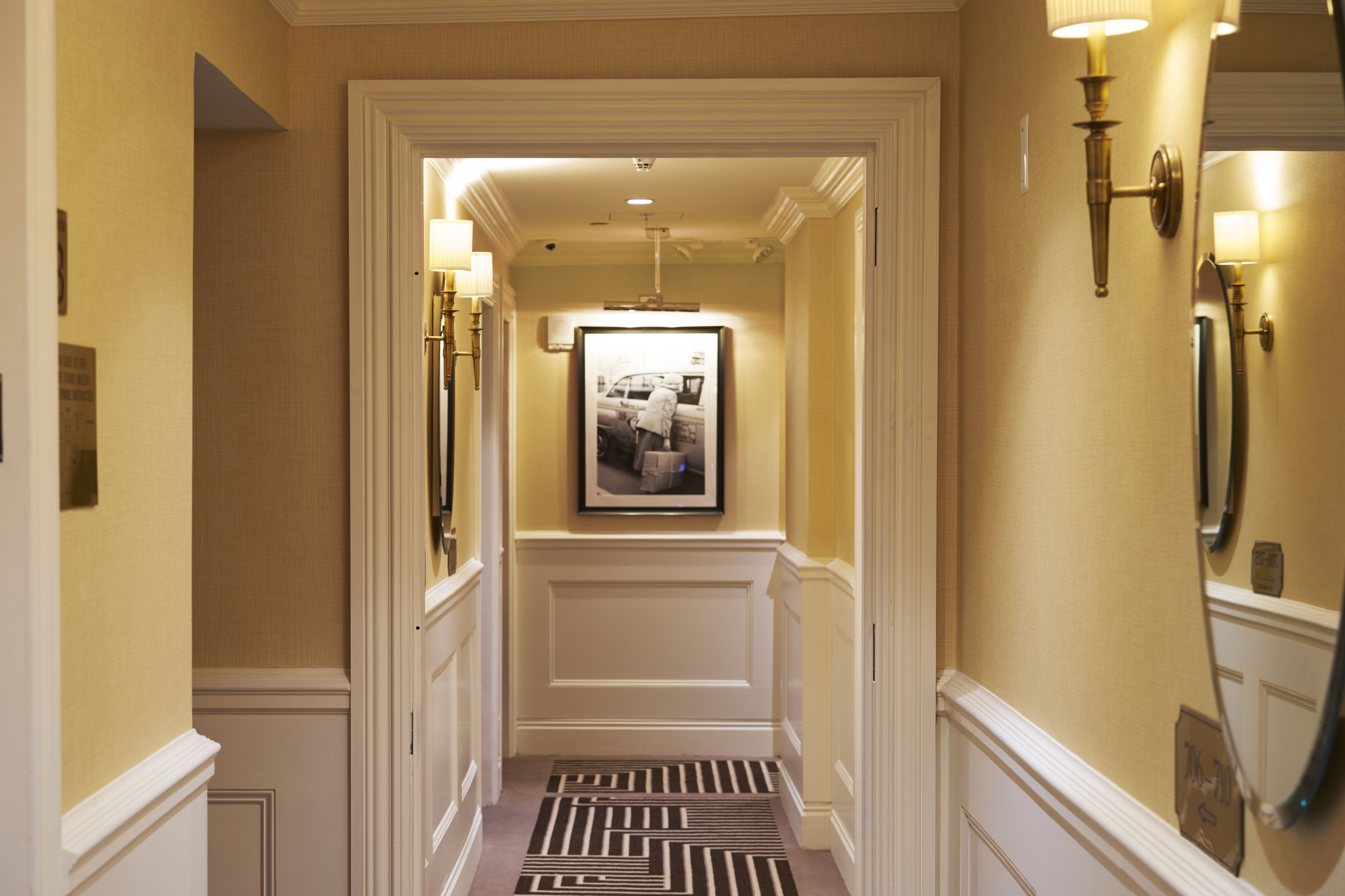 A hallway with a patterned carpet, a framed picture on the wall, and wall sconces.