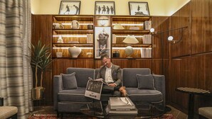 A man sitting on a grey sofa in a room with wooden paneling, a bookshelf, and a patterned rug. - The Iroquois Times Square, a Small Luxury Hotel (New York)