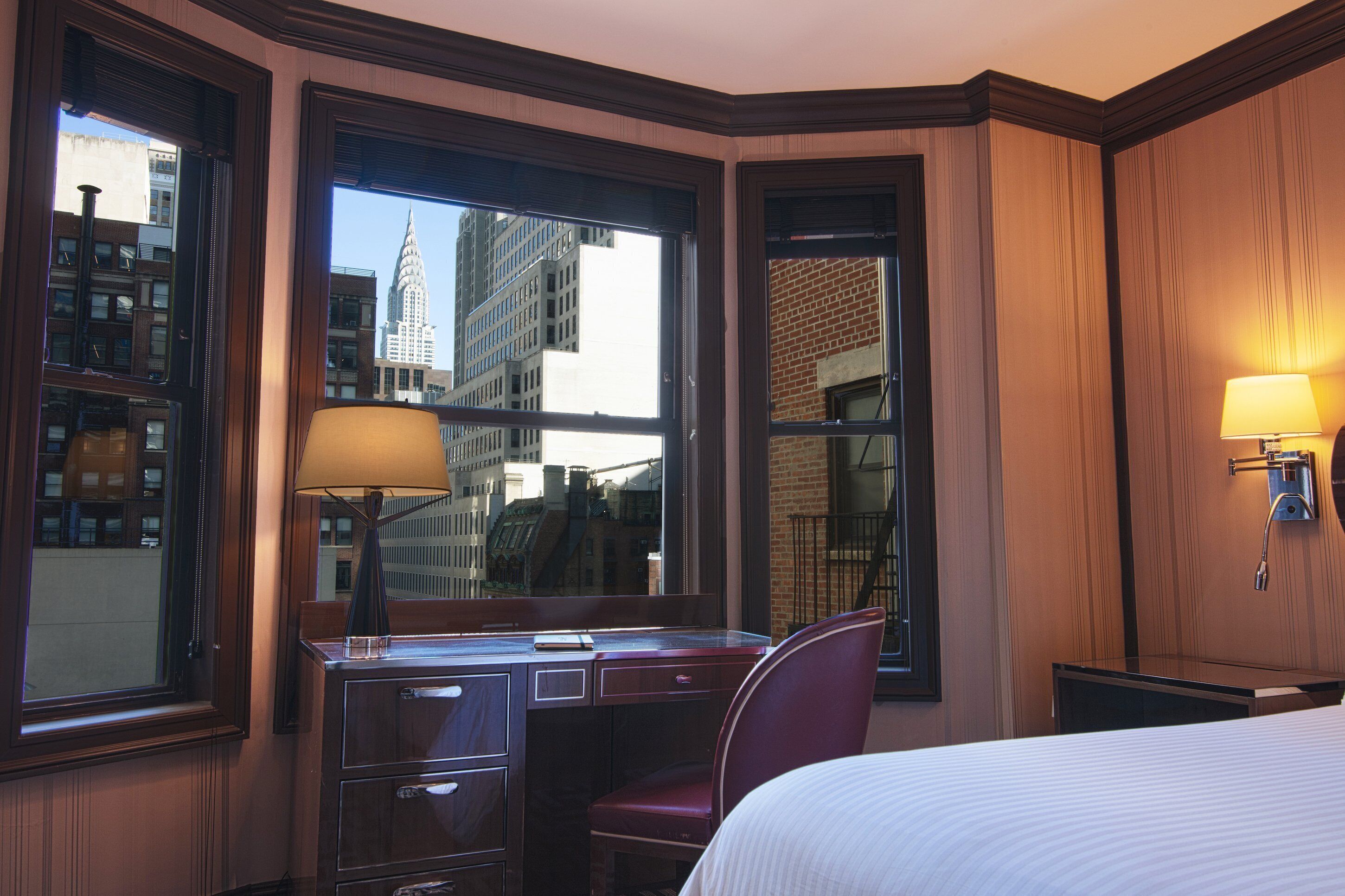 Een hotelkamer met een bureau, stoel en bed. Grote ramen bieden uitzicht op de skyline.