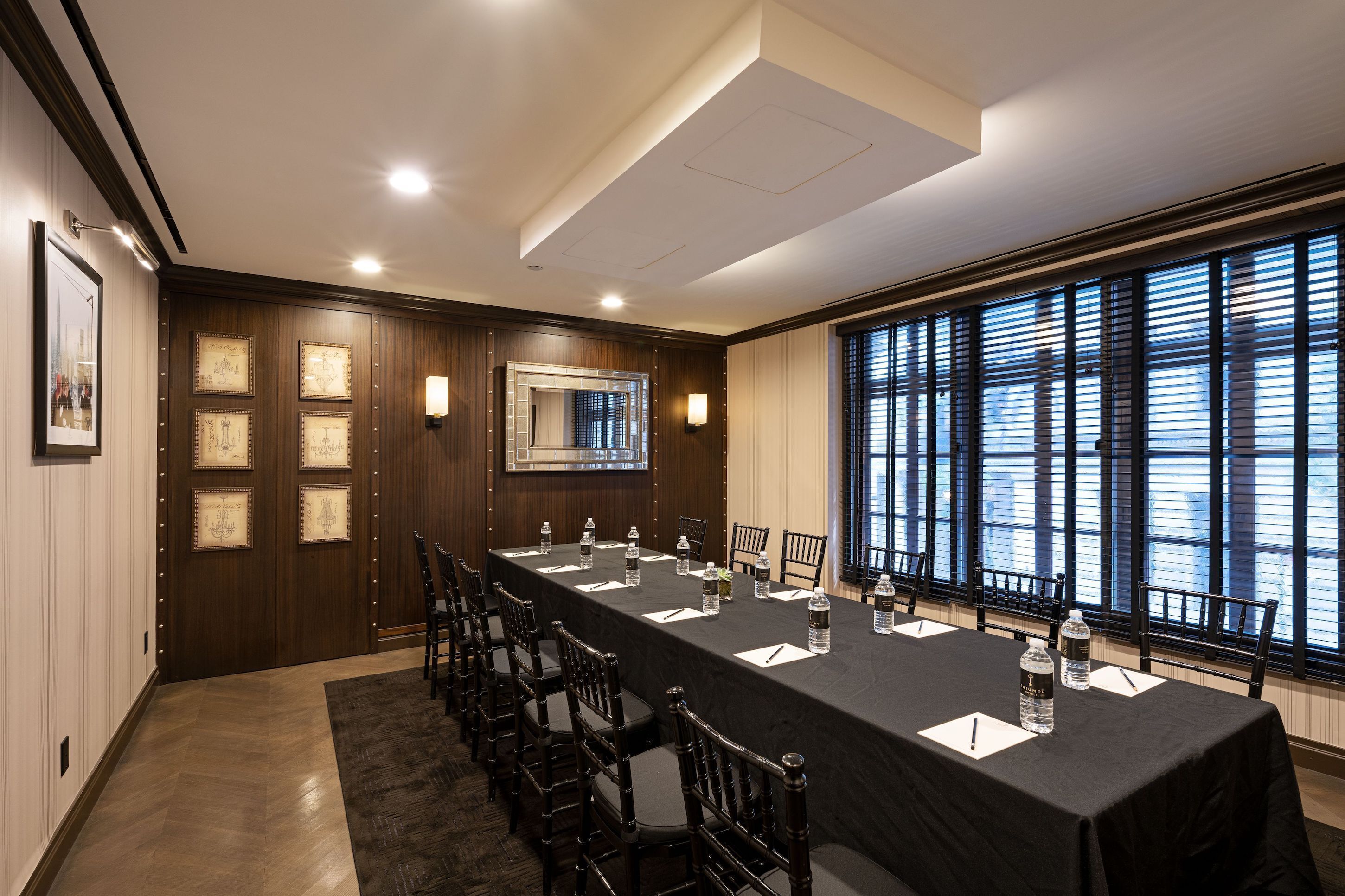 Sala de conferência com uma mesa comprida, cadeiras, quadros e uma grande janela com persianas.