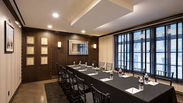 Sala de conferência com uma mesa comprida, cadeiras, quadros e uma grande janela com persianas.