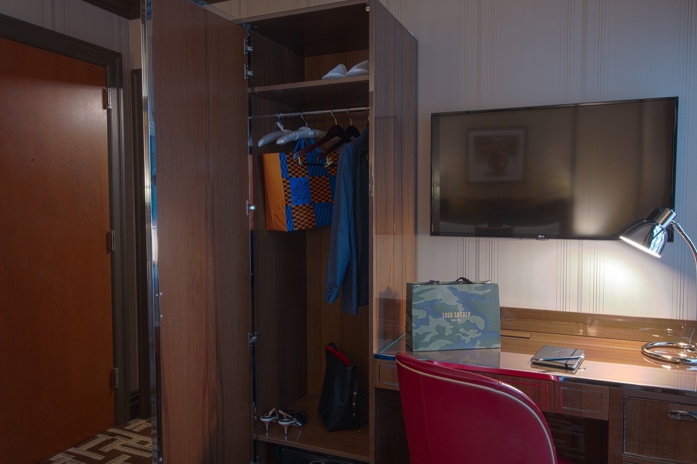A hotel room with a wooden wardrobe, a desk with a laptop, a red chair, and a television on the wall.