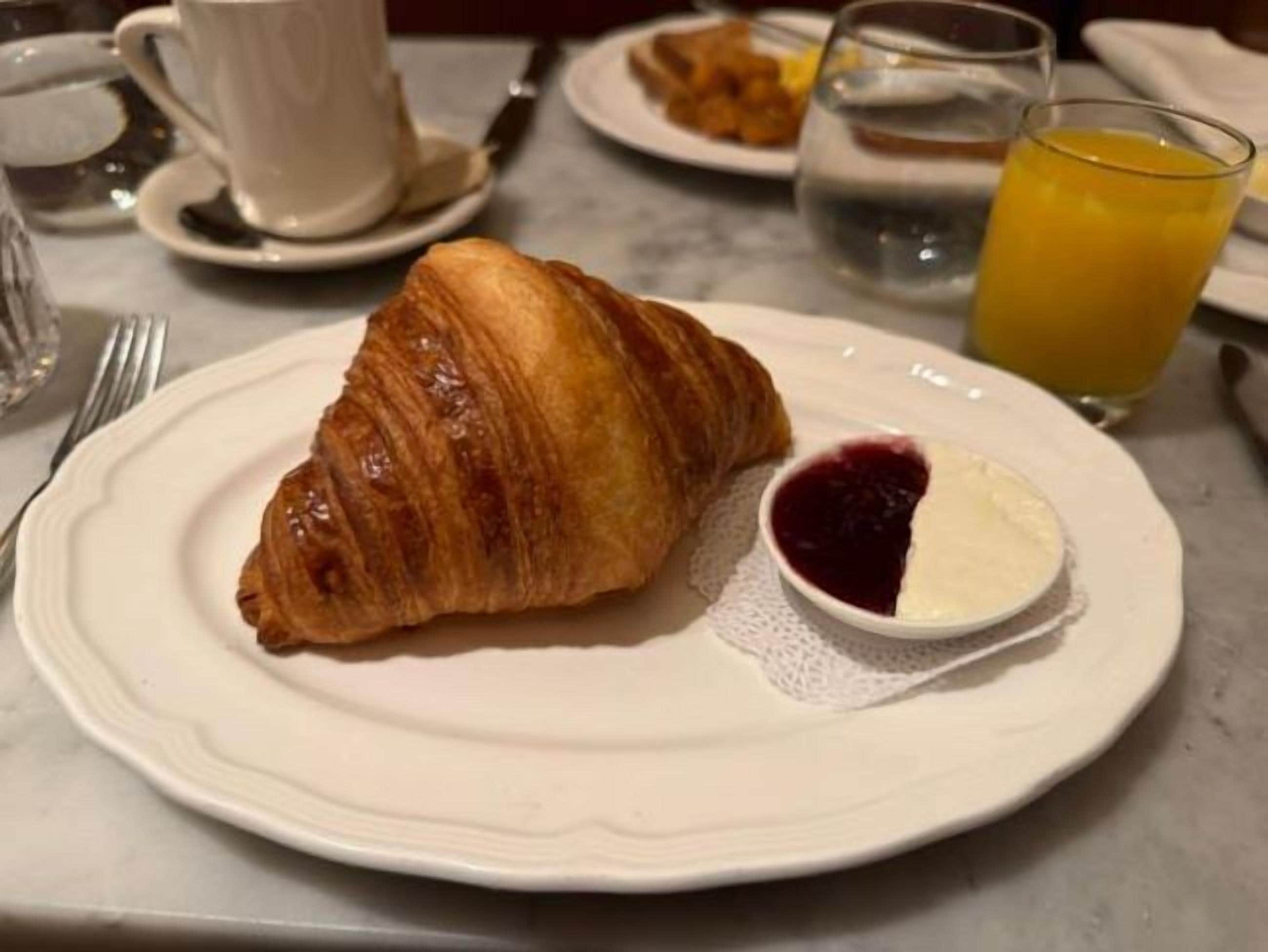 Un croissant sobre un plato con un recipiente pequeño de mermelada y un vaso de zumo de naranja.