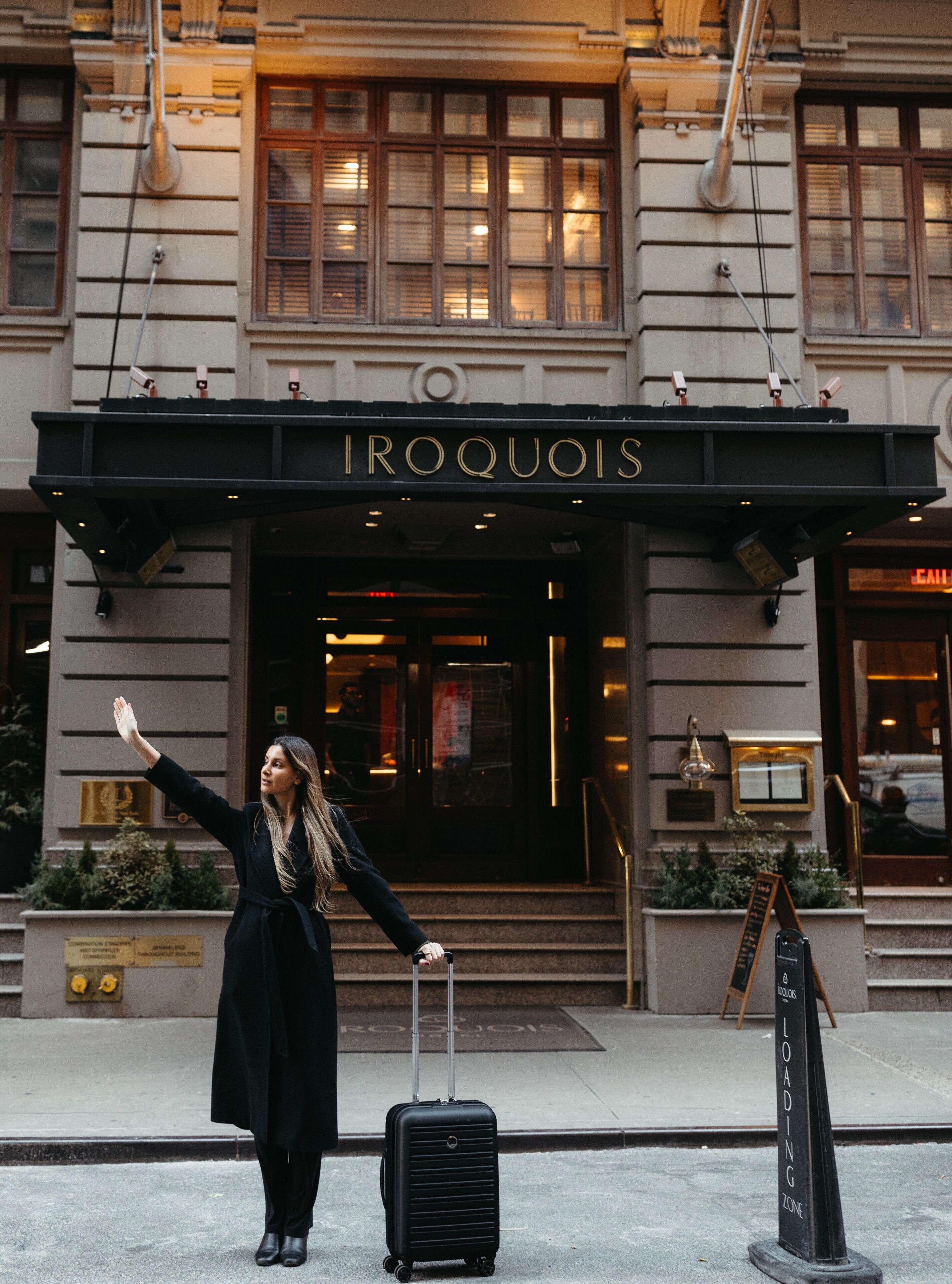 Une femme se tient devant un bâtiment sur lequel on peut lire l’inscription « IROQUOIS ». Elle tient une valise et fait signe de la main.