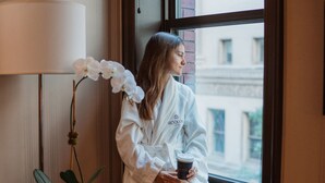 A person in a white robe sitting by a window with a view of buildings outside.