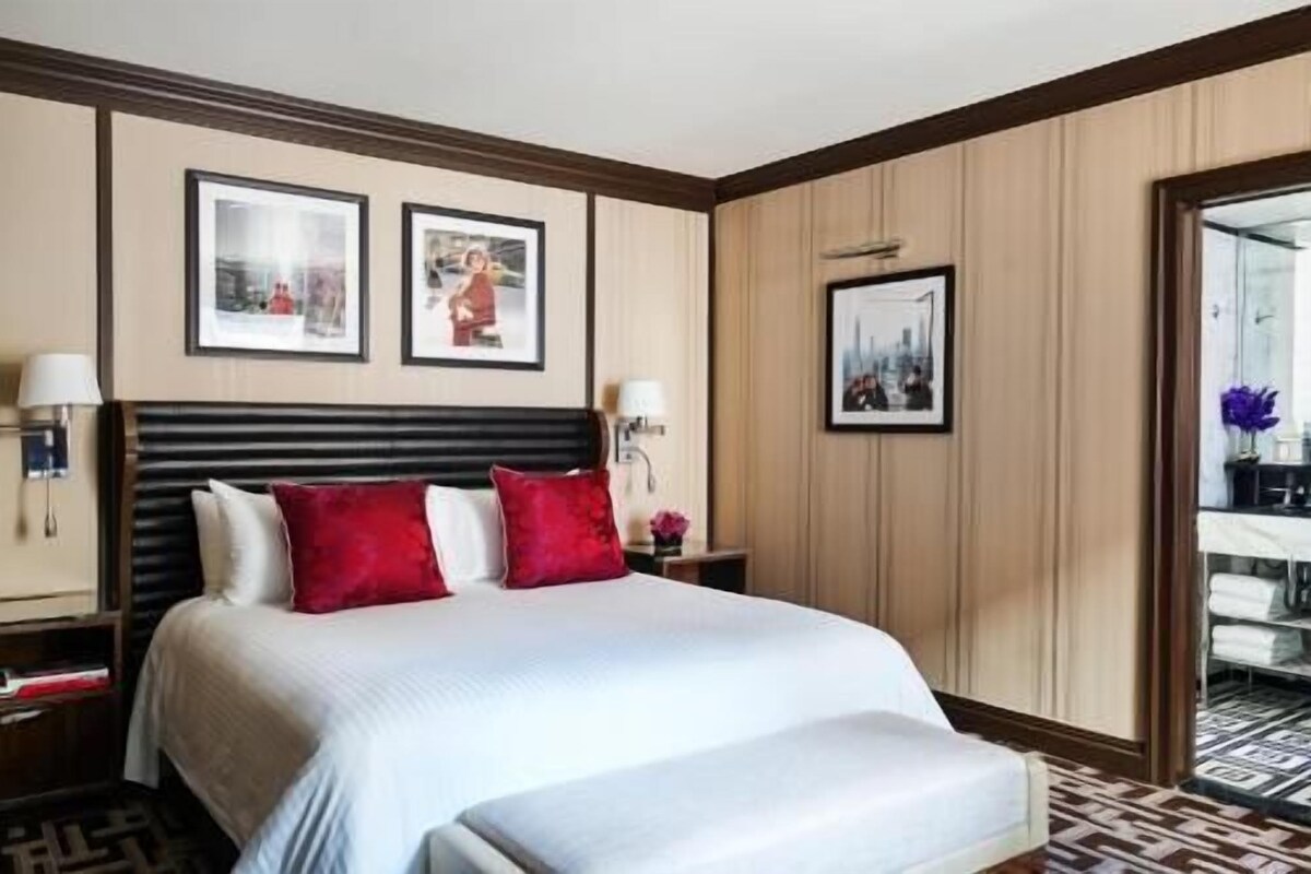 Habitación de hotel con una cama grande, dos cuadros en la pared, una mesita de noche con lámpara y vistas al exterior a través de una puerta de cristal.