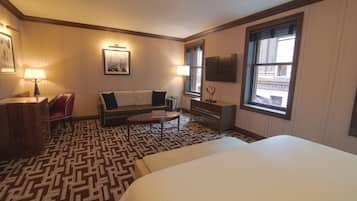 A hotel room with a large bed, a sofa, a small table, a desk, and a window with a view of buildings.
