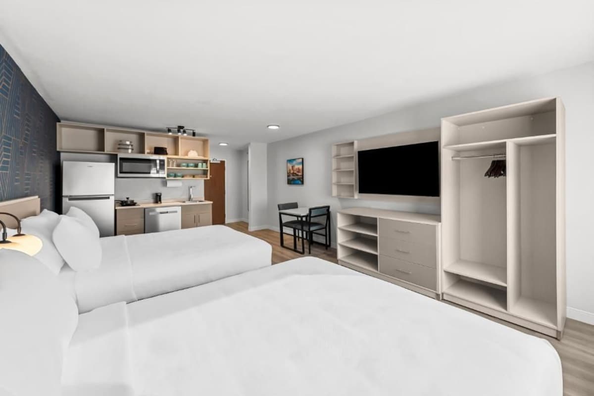 A modern hotel room with a bed, kitchenette, TV, and wardrobe.