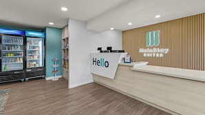 Et hotellets receptionsområde med et 'Hello'-skilt, en receptionsdisk, en væg beklædt med træpaneler og et køleskab fyldt med drikkevarer.