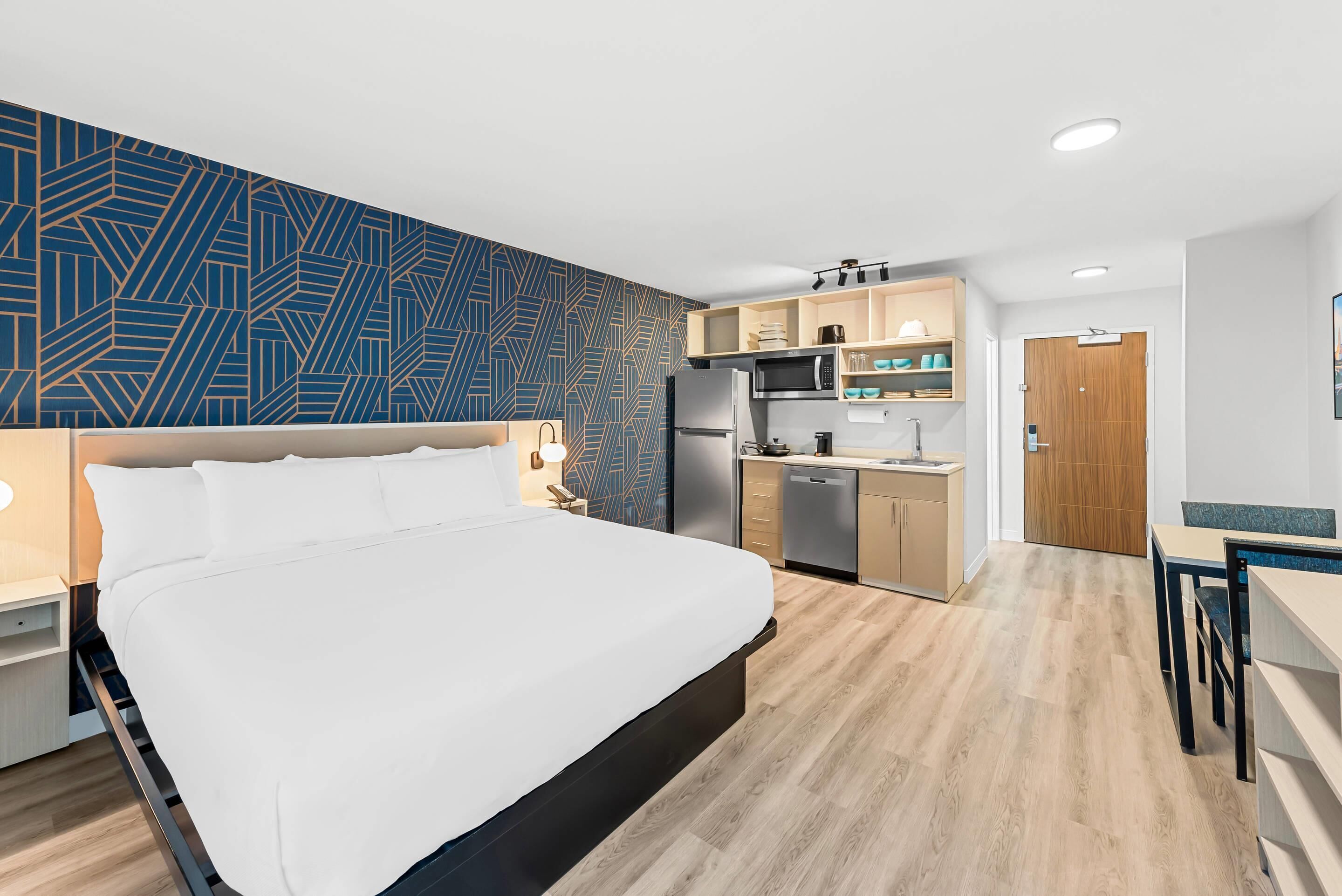 A modern hotel room with a large bed, a kitchenette, and a dining area.