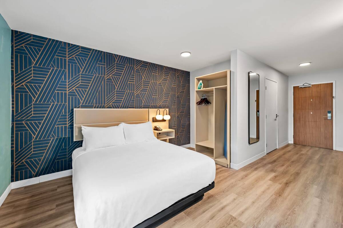 A modern hotel room with a large bed, a geometric-patterned wall, a wooden floor, a walk-in closet, and a mirror.