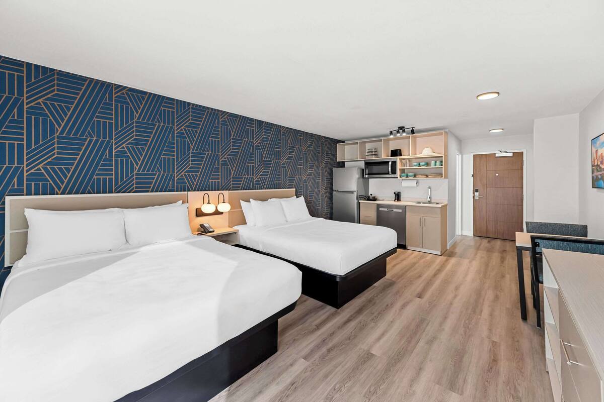 A modern hotel room with two beds, a kitchenette, and a dining area.
