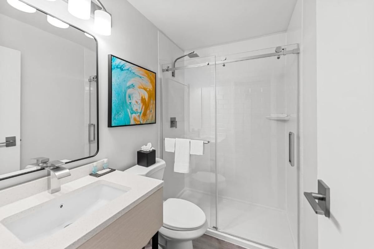 A modern bathroom with a large mirror, two sinks, a toilet, and a glass-enclosed shower.