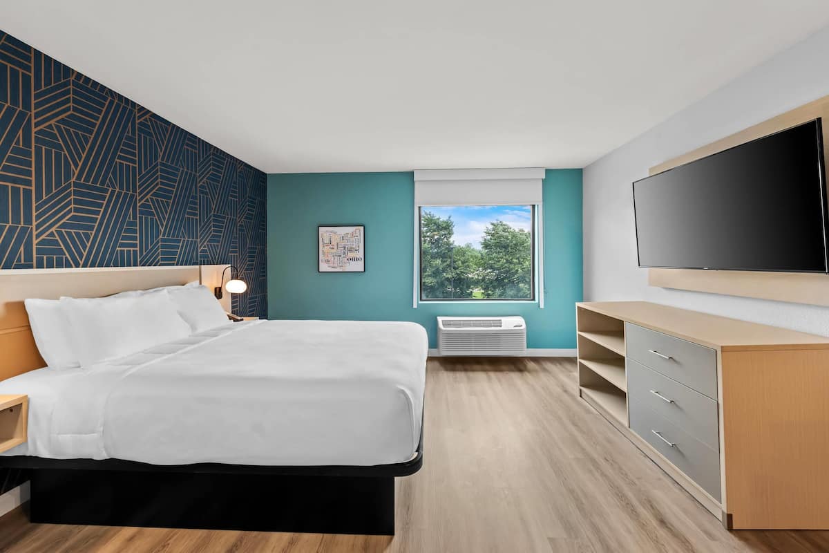 A modern hotel room with a bed, a flat-screen TV, a desk, and a window with a view of trees.