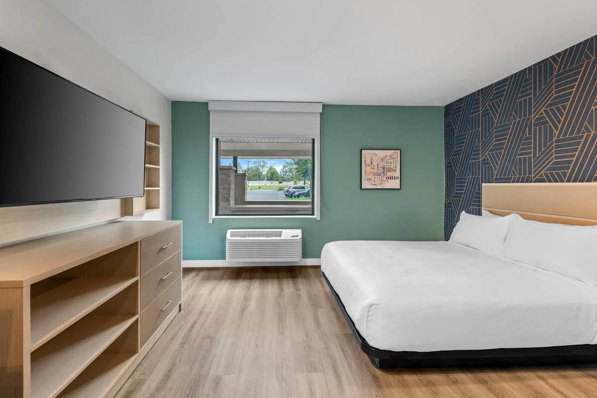 A modern hotel room with a large bed, a flat-screen TV, a wooden dresser, and a window with a view of the parking lot.