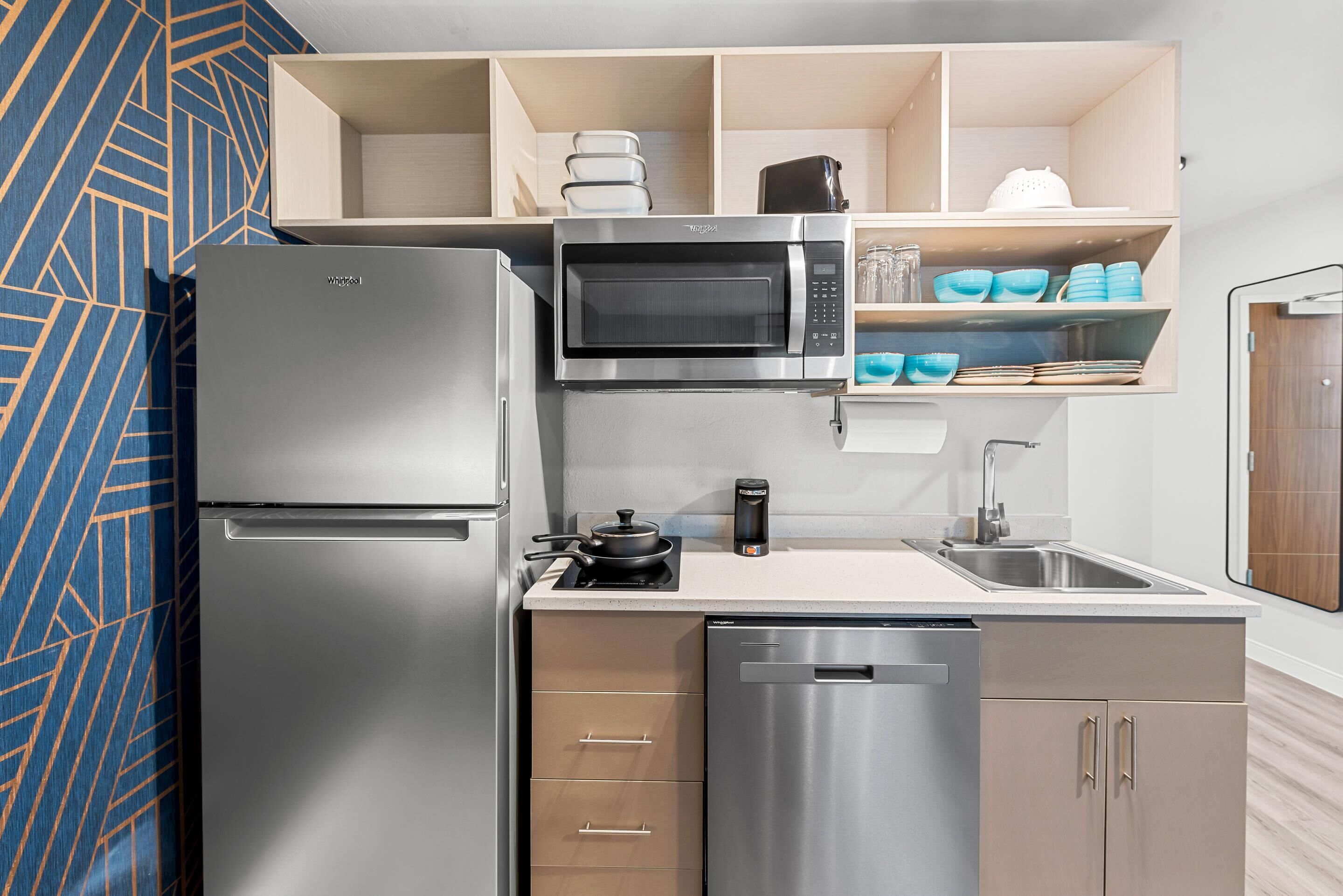 A compact kitchen with a stainless steel refrigerator, microwave, and dishwasher. There are wooden cabinets with a built-in shelf above the sink, and a patterned wall to the left.