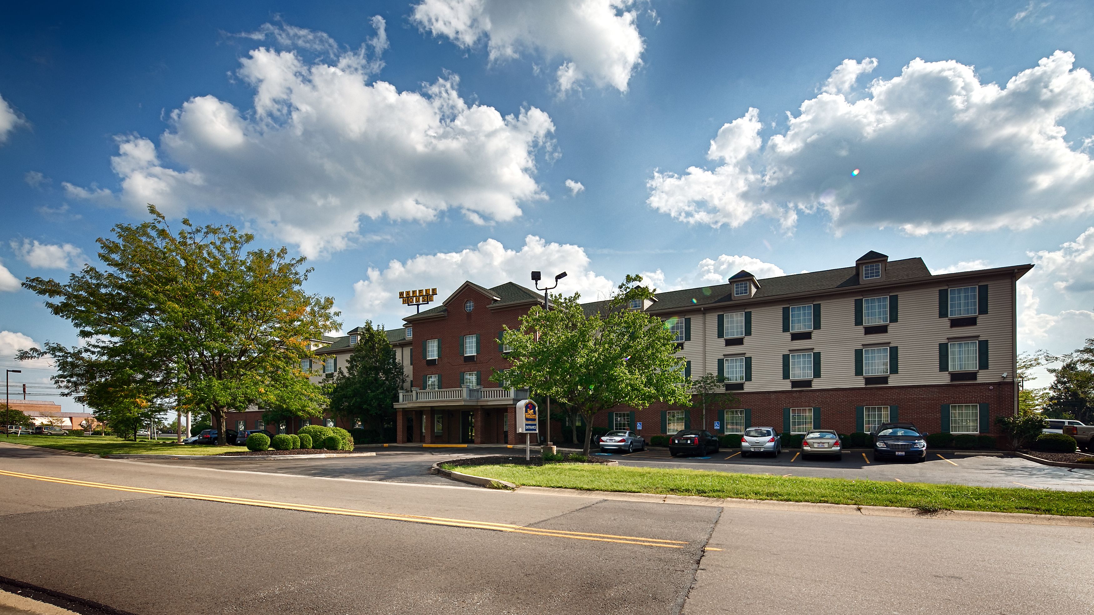 A multi-story hotel with a brick facade, a visible entrance, and a parking lot with several cars.