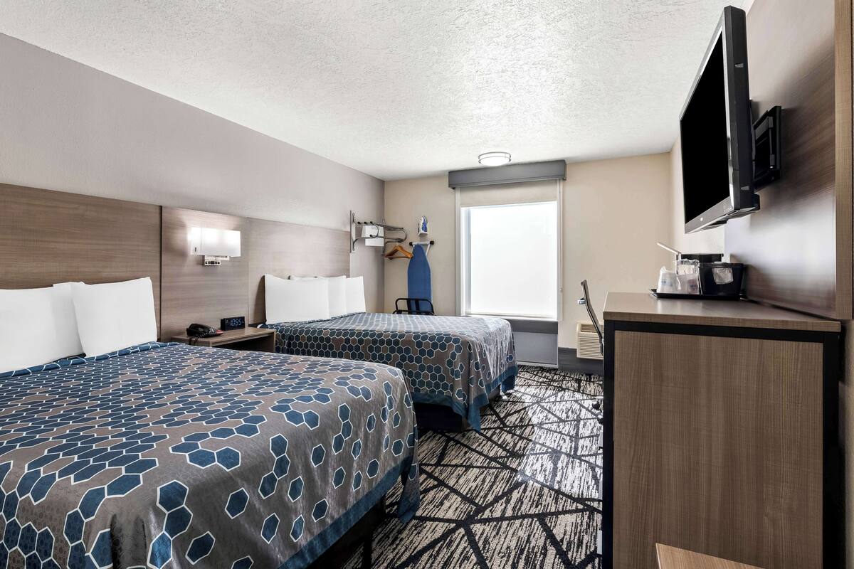 A hotel room with two beds, a desk, a TV, a window, and a wall-mounted TV.