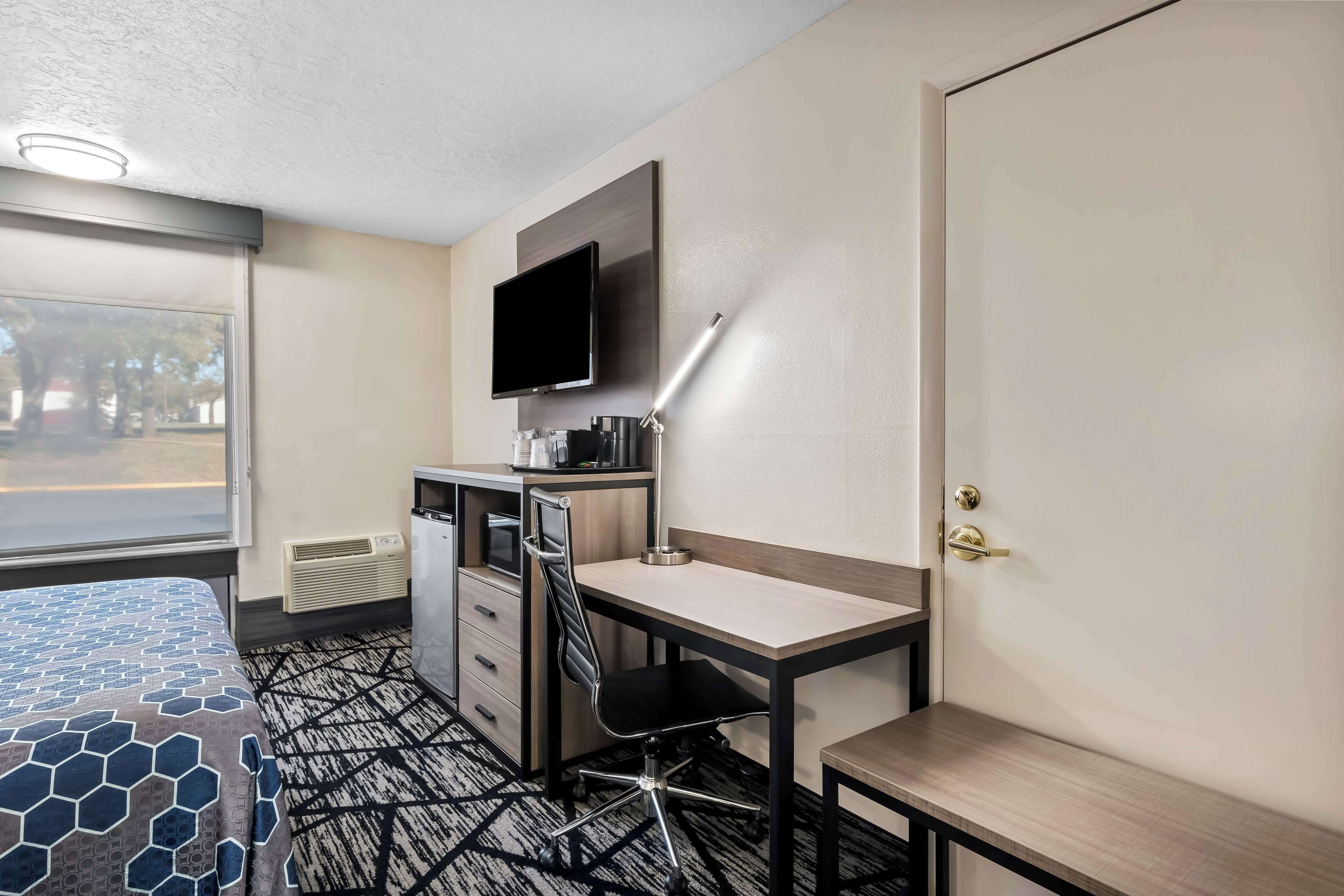 Quarto de hotel com cama, escrivaninha, cadeira, televisão e janela.