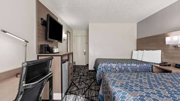 Habitación de hotel con escritorio, silla, cama con ropa de cama estampada, televisor y mesita de noche con lámpara.
