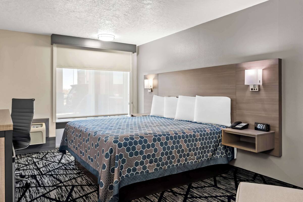 A hotel room with a bed, bedside table, lamp, and a view of the outside.