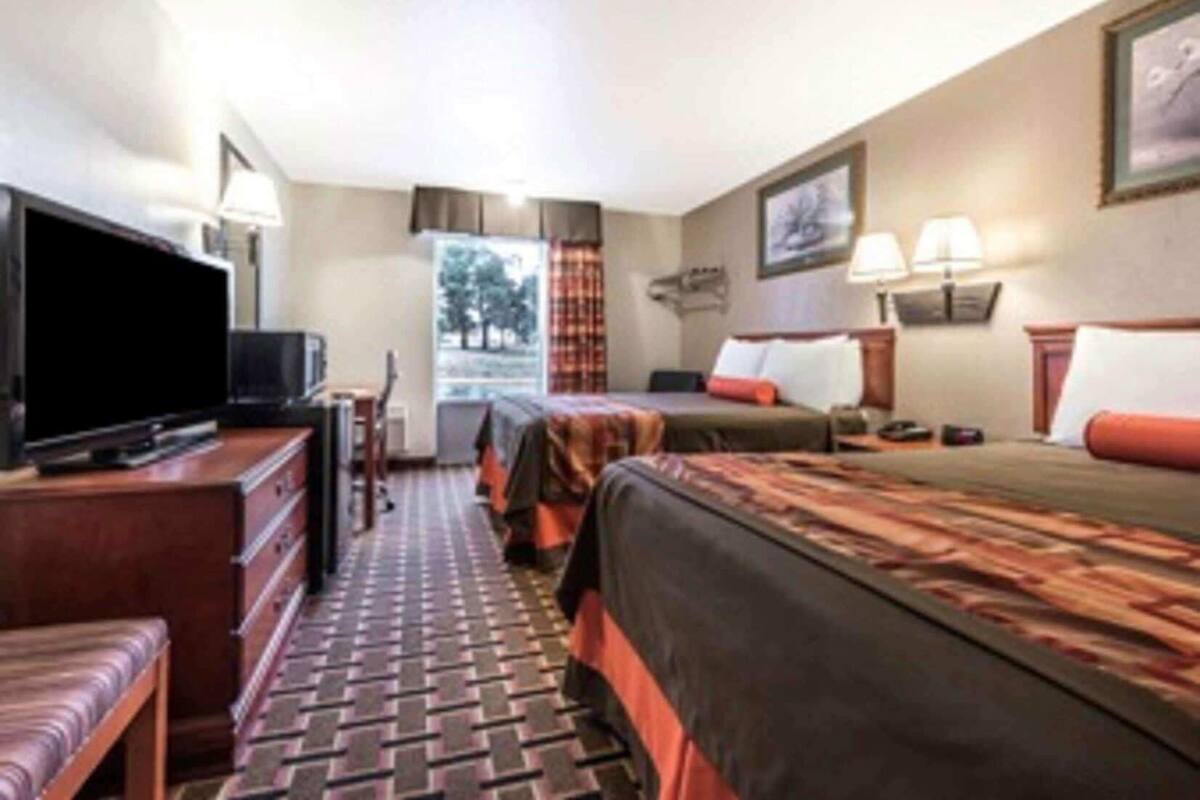 A hotel room with a television, a desk, a chair, a bed, a window with curtains, and a wall-mounted lamp.
