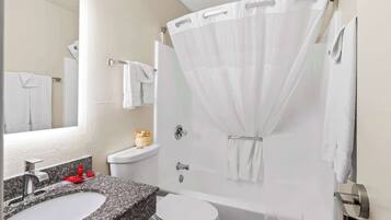 A bathroom with a granite countertop sink, a white toilet, a bathtub with a white shower curtain, and a mirror above the sink.