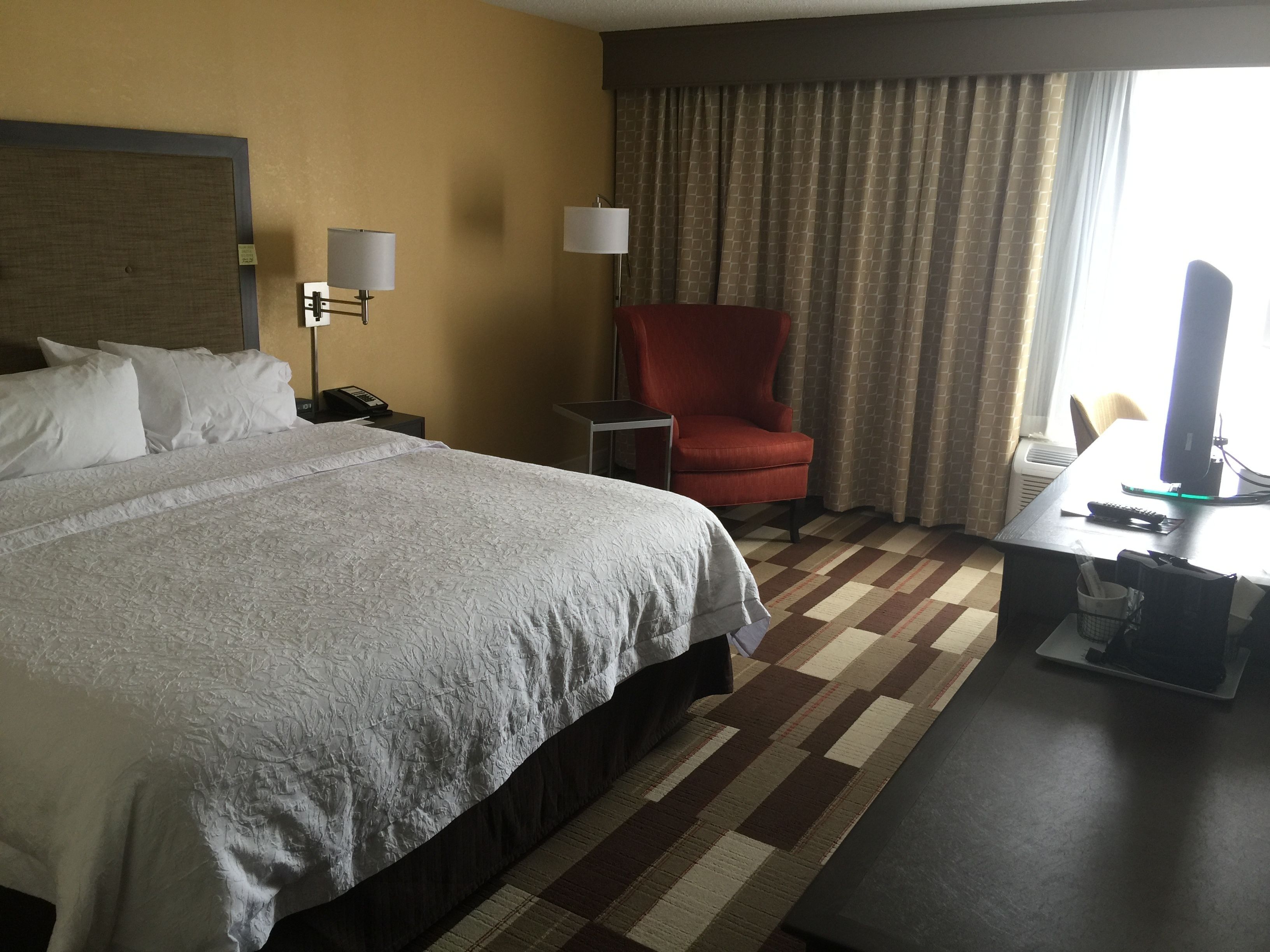 A hotel room with a large bed, a desk with a computer, a red armchair, and a window with curtains.